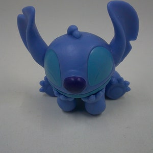 Puede incluir: Una figura azul de Stitch con ojos grandes y una nariz morada. La figura est&aacute; sentada con los brazos cruzados.