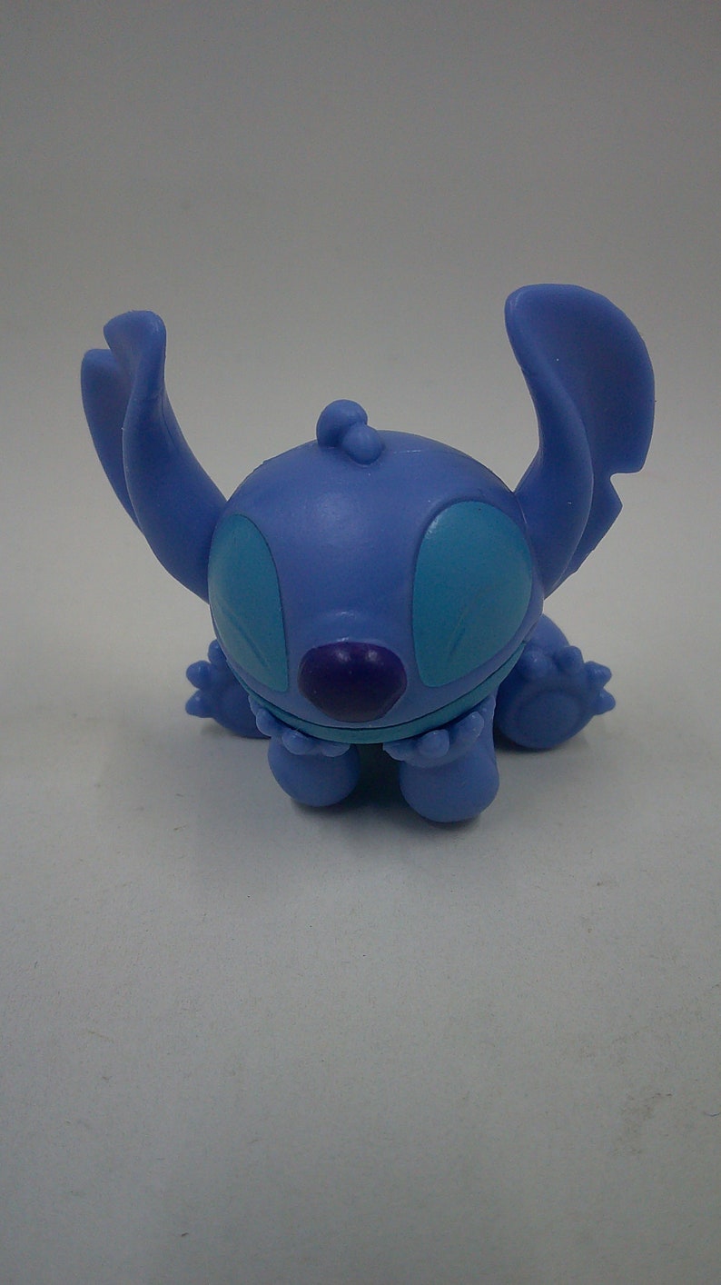 Puede incluir: Una figura azul de Stitch con ojos grandes y una nariz morada. La figura est&aacute; sentada con los brazos cruzados.