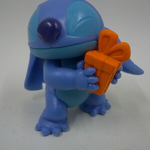 Puede incluir: Personaje azul Stitch sosteniendo una caja de regalo naranja brillante con un lazo. El personaje tiene un cuerpo azul, ojos azules y una nariz morada.