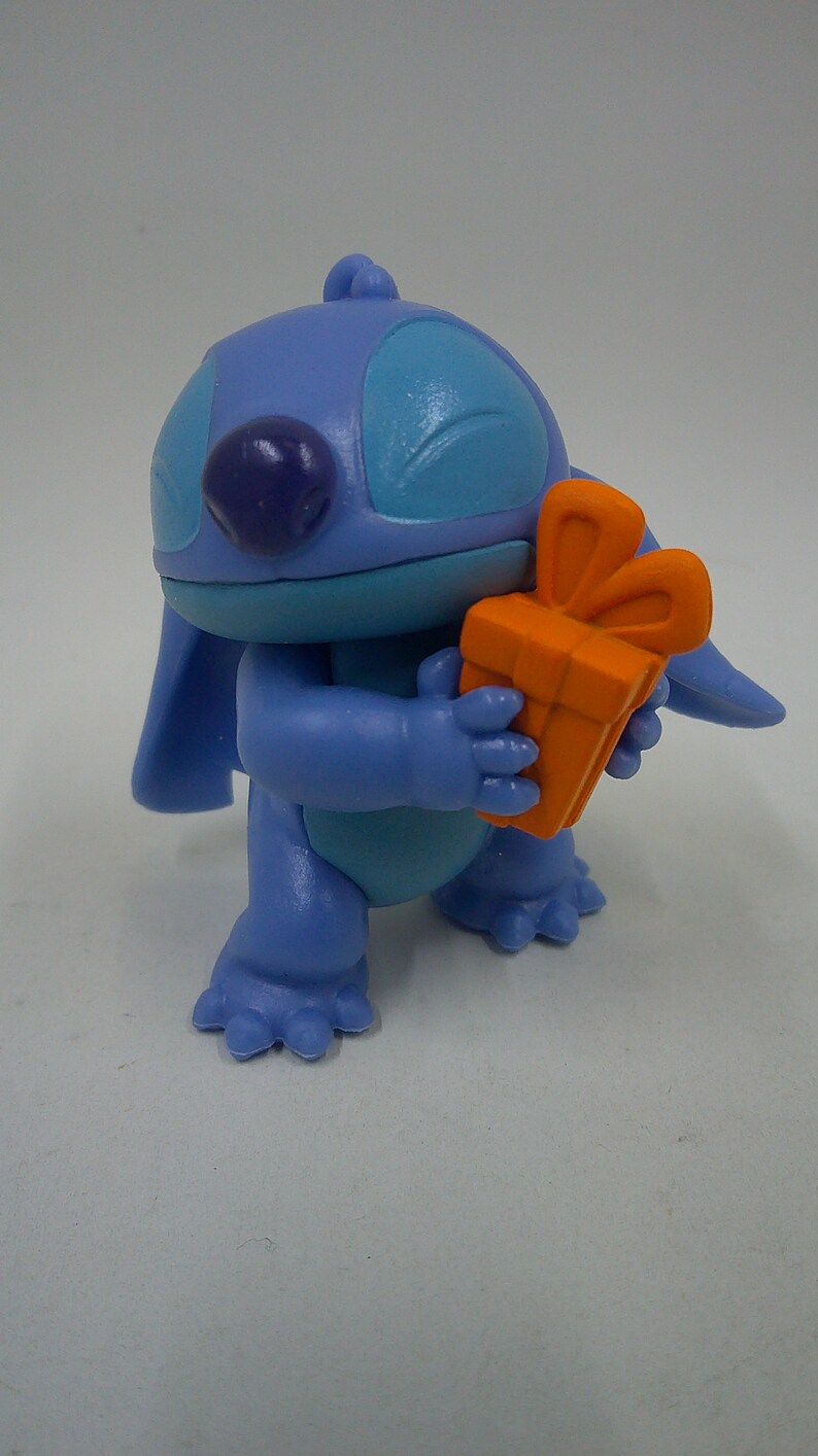 Puede incluir: Personaje azul Stitch sosteniendo una caja de regalo naranja brillante con un lazo. El personaje tiene un cuerpo azul, ojos azules y una nariz morada.