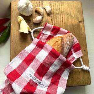 May include: A red and white checkered bread bag with a loaf of bread inside, sitting on a wooden cutting board. The bag has a drawstring closure and a label that reads "PORTER ARBON". Also on the board are garlic cloves, butter, and chili peppers.