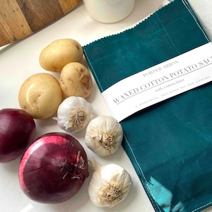 May include: A teal waxed cotton potato sack is pictured with fresh produce. The produce includes yellow potatoes, red onions, and garlic bulbs. The sack has a white label with the text "WAXED COTTON POTATO SACK".