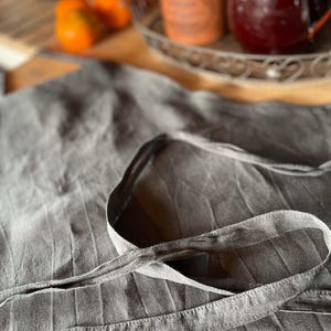 Puede incluir: Un delantal de lino gris con una tira larga atada. El delantal está hecho de una tela texturizada con un sutil patrón a rayas.