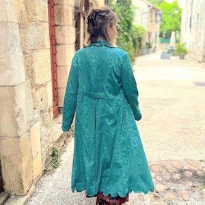 May include: A teal-colored long coat with a floral pattern and a scalloped hemline. The coat has long sleeves and a belt at the waist. The person is walking down a cobblestone street.