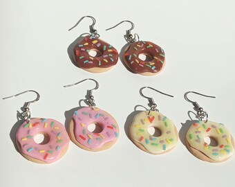 May include: Six pairs of donut-shaped earrings with silver hooks. Two pairs have chocolate frosting and colorful sprinkles. Two pairs have pink frosting and sprinkles. The remaining two pairs have yellow frosting and sprinkles.