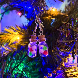Op de afbeelding: Een paar unieke oorbellen met miniatuur glazen flesjes met kurken stoppen. Elke fles bevat kleurrijke, snoepachtige stukjes in rood, groen en wit. De oorbellen hangen aan zilverkleurige haken, perfect voor een grillige touch.