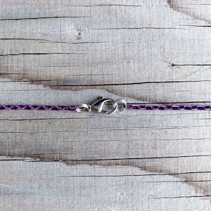 May include: Close-up of a purple chain necklace with a silver-colored clasp. The chain is made of small, linked segments, and the clasp is a lobster claw style. The background is a weathered, light-colored wooden surface.