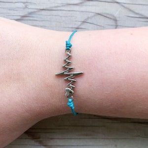 Op de afbeelding: Een turquoise armband van touw met een zilveren hartslagbedel.