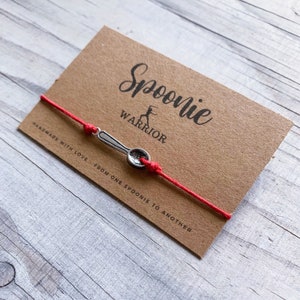 May include: A red string bracelet with a small silver spoon charm. The bracelet is on a brown card with the words "Spoonie Warrior" and the phrase "Handmade with love from one spoonie to another."