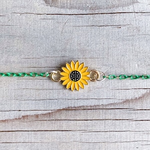 May include: A sunflower charm bracelet. The bracelet features a yellow and black sunflower pendant with a gold-colored chain and green chain links. The bracelet is set against a weathered, light-colored wooden surface.