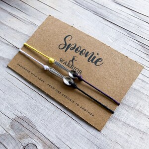 Könnte beinhalten: Drei Löffelarmbänder mit silbernen Löffelanhängern und verstellbaren Kordeln in Gelb, Weiß, Schwarz und Lila. Die Armbänder werden auf einer braunen Karte mit dem Text "Spoonie Warrior" und "Handmade with love..." präsentiert.