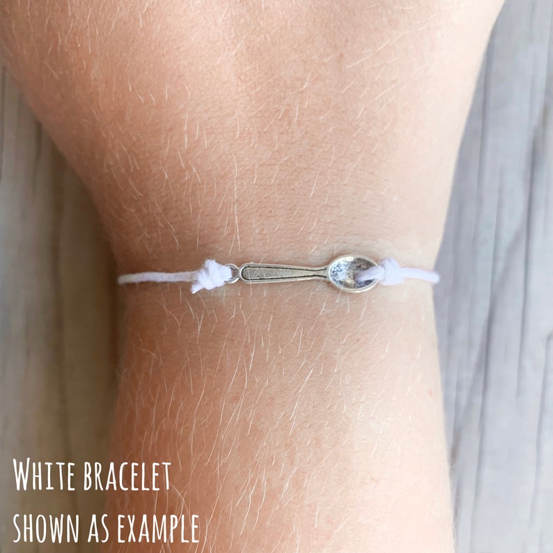 May include: White bracelet with a silver spoon charm.  "White Bracelet Shown as Example" is written below the bracelet.