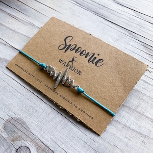 May include: Teal string bracelet with a silver heart beat charm and the words "Spoonie Warrior" on a brown card.