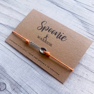 Könnte beinhalten: Ein Armband mit einem silbernen Löffelanhänger und einer orangefarbenen Kordel, präsentiert auf einer braunen Karte. Die Karte trägt die Worte "Spoonie Warrior" und den Satz "Handmade with love - From one spoonie to another."