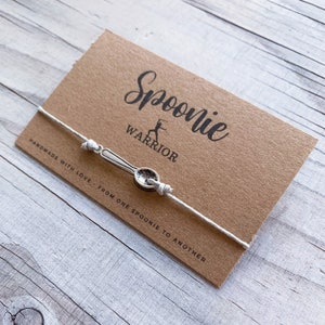 May include: A white string bracelet with a silver spoon charm. The bracelet is on a brown card with the text "Spoonie Warrior" and "Handmade with love, from one spoonie to another."
