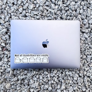 May include: A silver laptop with a sticker that reads "Not all disabilities are visible." The sticker features a line of five cartoon ghosts.