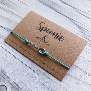 May include: A teal cord bracelet with a silver spoon charm. The bracelet is displayed on a brown card with the words "Spoonie Warrior" and "Handmade with love - From one spoonie to another."