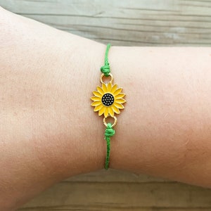 Puede incluir: Un brazalete de cuerda verde con un dije de girasol dorado. El girasol es amarillo con un centro negro.