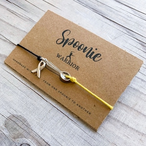 Puede incluir: Una pulsera Spoonie Warrior con un dije de lazo amarillo y un dije de cuchara, sobre una tarjeta marrón. La tarjeta tiene las palabras "Spoonie Warrior" y "From one spoonie to another". La pulsera está hecha a mano.