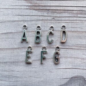 May include: Silver metal alphabet letter charms, A through G, on a light wood background.