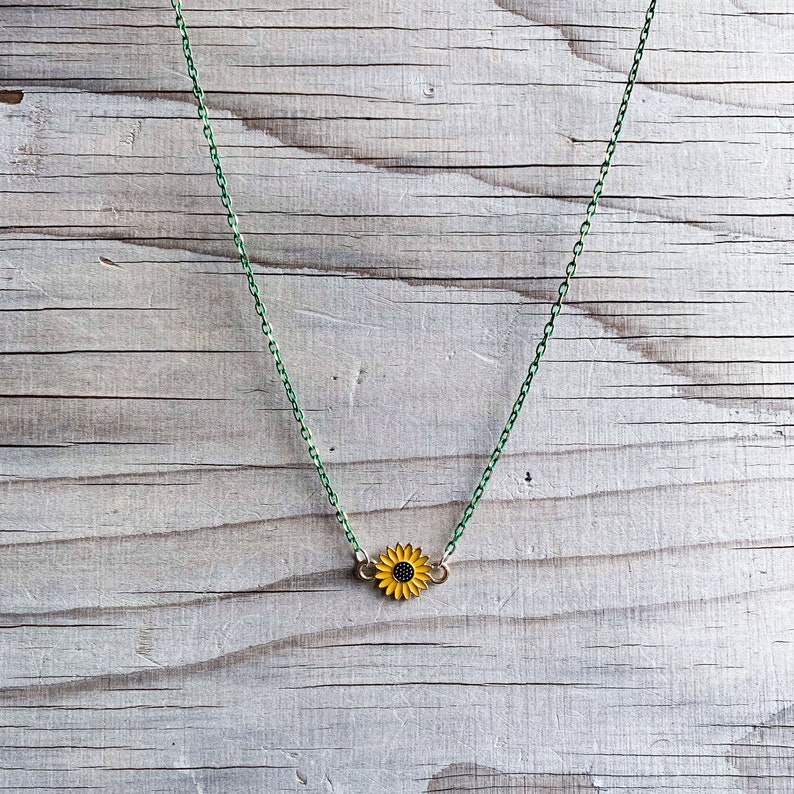 May include: A delicate necklace featuring a sunflower pendant. The pendant has yellow petals and a black center. The chain is a vibrant green color, creating a striking contrast against the wooden background.