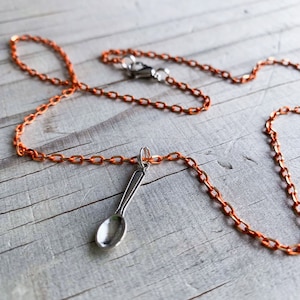 May include: A silver spoon charm hangs from a copper chain necklace. The chain is made of small, linked circles.
