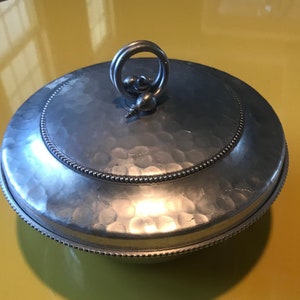 Könnte beinhalten: Ein silberfarbener, runder Behälter mit Deckel und gehammerter Textur. Der Deckel hat einen Perlenrand und einen Ringgriff mit einer kleinen dekorativen Kugel. Der Behälter steht auf einer gelben Oberfläche.