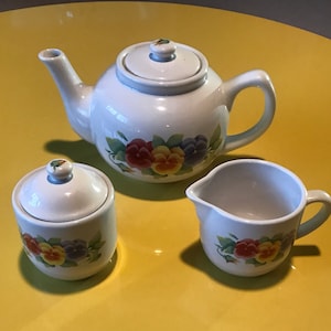 Op de afbeelding: Een witte theepot, suikerpot en roomkan set met een bloemenpatroon van gele, rode en paarse viooltjes. De set staat op een geel oppervlak.