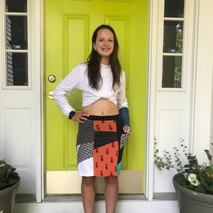 May include: A young person wearing a white long-sleeved shirt tied at the waist, a colourful patchwork skirt with a pineapple print, and a blue cast on their arm. They are standing in front of a green door with a brown doormat.