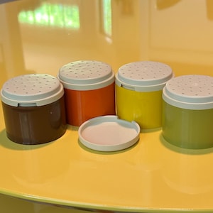 Può includere: Un set di quattro shaker per spezie colorati con coperchi bianchi e un piccolo coperchio rotondo bianco. Gli shaker sono marroni, arancioni, gialli e verdi. Gli shaker sono cilindrici e hanno piccoli fori sulla parte superiore.