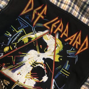 May include: Black t-shirt with a yellow and red "Def Leppard" logo and a graphic of a screaming face in a triangle. The text "Tour 1987" is visible at the bottom of the graphic.