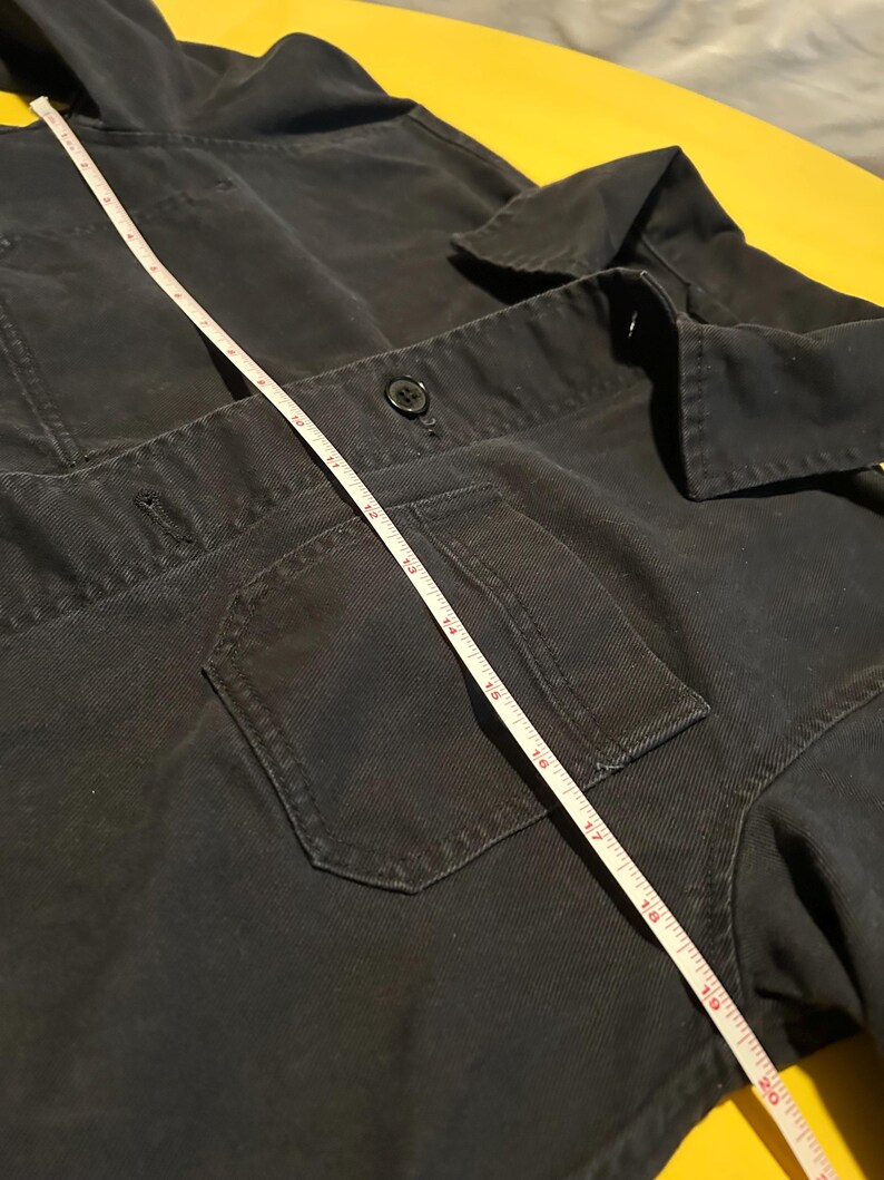 May include: Dark navy blue work jacket with a hood, two front pockets, and a button closure. A white measuring tape is laid across the jacket, showing a measurement of approximately 20 inches.