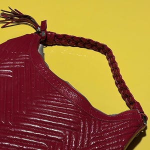 Puede incluir: Un bolso de mano de cuero acolchado rojo con un asa trenzada y un detalle de borla. El bolso tiene un cierre de cremallera y un acabado brillante. El diseño del bolso presenta un patrón de chevrón.