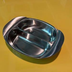 Puede incluir: Una bandeja de servicio dividida de acero inoxidable con acabado cepillado. La bandeja ovalada está dividida en dos compartimentos por un divisor central. La bandeja tiene bordes curvos y una superficie brillante y reflectante.