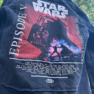 May include: Black denim jacket featuring a Star Wars graphic on the back. The design includes the Star Wars logo, Darth Vader, and text that reads "Episode V" and "The Empire Strikes Back."