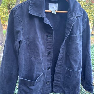 May include: A dark navy blue jacket with four front pockets and a button-up closure. The jacket is hanging on a wooden hanger. The brand label "H&M" is visible on the collar.