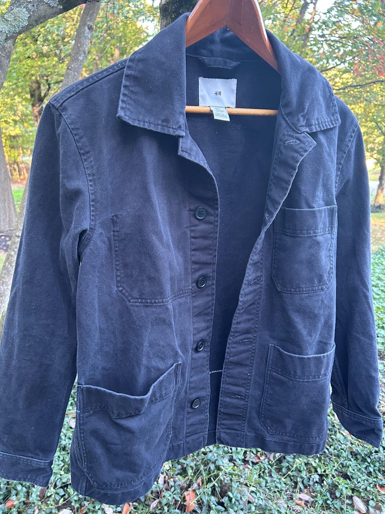 May include: A dark navy blue jacket with four front pockets and a button-up closure. The jacket is hanging on a wooden hanger. The brand label "H&M" is visible on the collar.