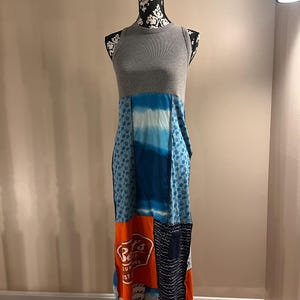 May include: A sleeveless maxi dress on a mannequin. The top is gray, transitioning to a patchwork skirt with blue, orange, and white panels. One panel features text. The dress is a unique, colorful garment.
