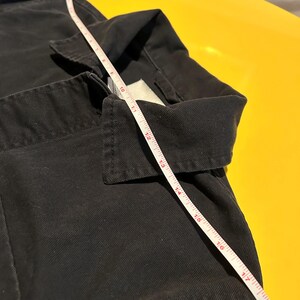 May include: Close-up of a black garment with a white measuring tape laid across it. The tape shows measurements in inches. The garment has a collar and appears to be made of a sturdy fabric. The background is a bright yellow.