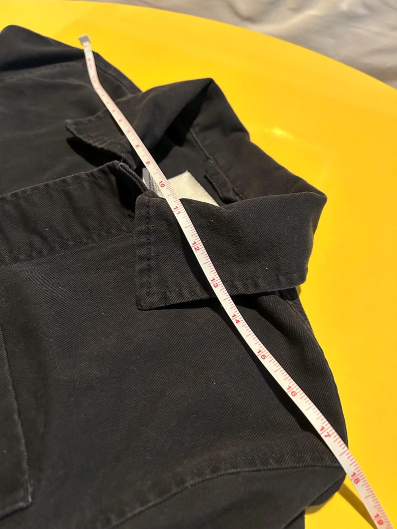 May include: Close-up of a black garment with a white measuring tape laid across it. The tape shows measurements in inches. The garment has a collar and appears to be made of a sturdy fabric. The background is a bright yellow.