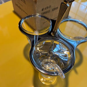 Könnte beinhalten: Ein Chrom- und Glas-Karussell-Servierer mit drei durchsichtigen Glasschalen und einem Griff. Der Servierer steht auf einer gelben Oberfläche, im Hintergrund eine Pappschachtel. Auf der Schachtel steht "Kromex" und "Carousel Server".