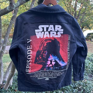 May include: Black denim jacket featuring a Star Wars graphic on the back. The graphic includes Darth Vader's image, the Star Wars logo, and text that reads "Episode V The Empire Strikes Back."
