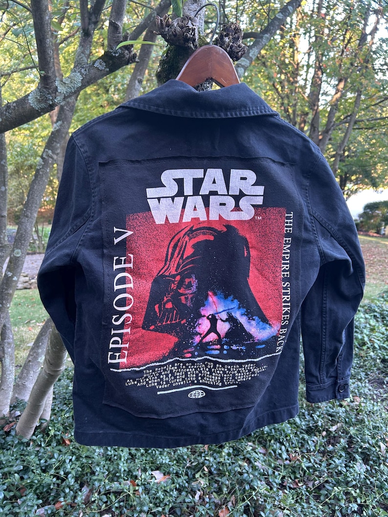 May include: Black denim jacket featuring a Star Wars graphic on the back. The graphic includes Darth Vader's image, the Star Wars logo, and text that reads "Episode V The Empire Strikes Back."