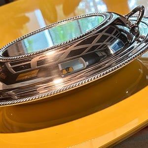 May include: An oval silver-toned serving dish with a polished, reflective surface and a domed lid. The dish features a decorative rope-style border along the rim and two small, upright metal handles on opposite ends for easy lifting and transport.