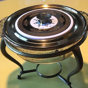Könnte beinhalten: Eine goldfarbene Metall-Chafing Dish mit einer Glasschale und einem Deckel mit einem weißen Porzellaneinsatz. Der Deckel hat einen goldfarbenen Knopf und einen goldfarbenen Rand. Der Porzellaneinsatz hat ein schwarzes und goldenes Design mit den Worten "Briard" und "Sevres" in Gold.