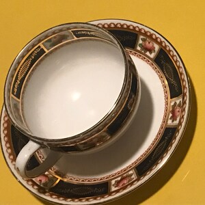 May include: A white teacup and saucer set with a decorative design. The cup and saucer have a dark blue border with gold trim and floral accents. The cup has a white interior.