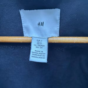 May include: Close-up of a navy blue garment tag with the H&M logo. The tag includes size information: EUR S, US S, CA P, CN 175/92A, MX CH. The tag also states "MADE IN BANGLADESH" and "FABRIQU&Eacute; AU BANGLADESH".