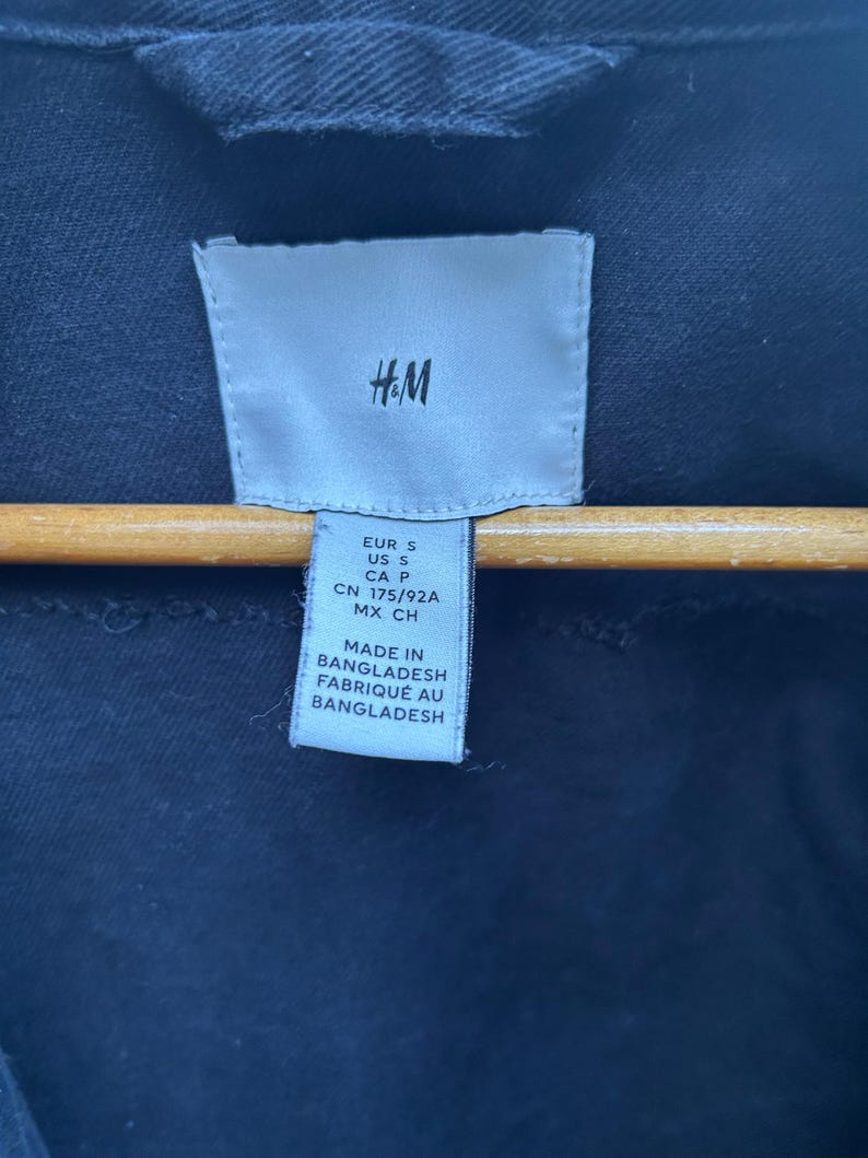 May include: Close-up of a navy blue garment tag with the H&M logo. The tag includes size information: EUR S, US S, CA P, CN 175/92A, MX CH. The tag also states "MADE IN BANGLADESH" and "FABRIQU&Eacute; AU BANGLADESH".