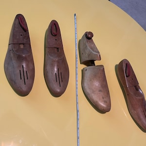 May include: Four wooden shoe forms in shades of brown. Each form has a red fabric detail at the top and vertical slits on the front. One form is split in two. A measuring tape is next to the forms.