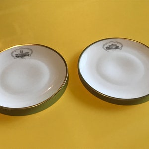 May include: Two white ceramic saucers with gold trim and a green underside. Each saucer features a black and white image of a building.